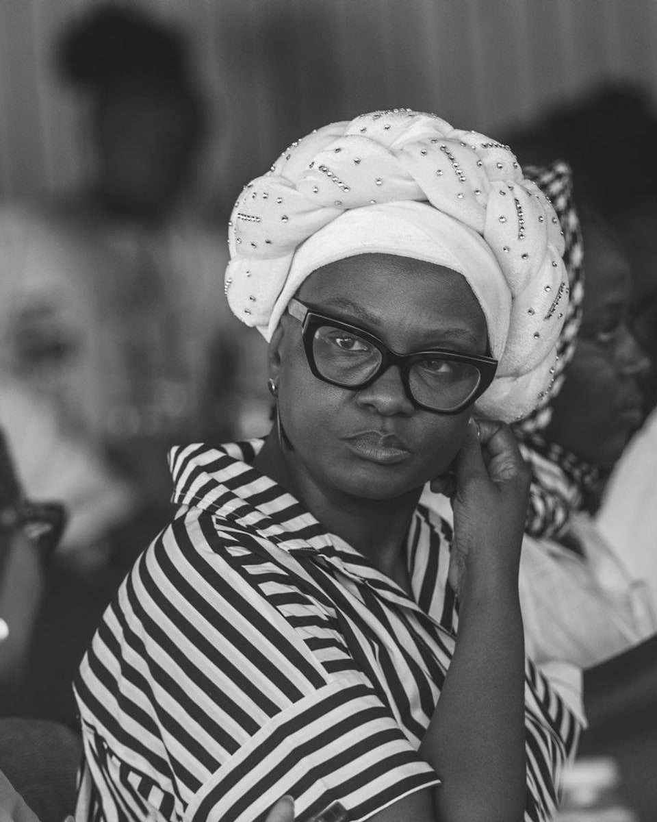 A woman in glasses and a white head wrap, resting her hand at her ear and listening with a questioning look.
