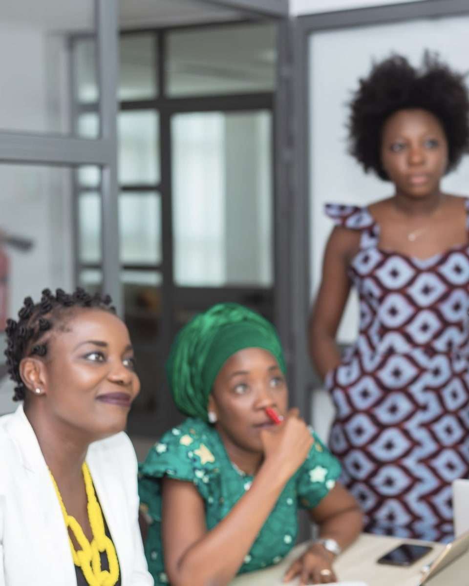 Women in an office meeting, one standing and the others seated, listening to a colleague who holds a tablet.